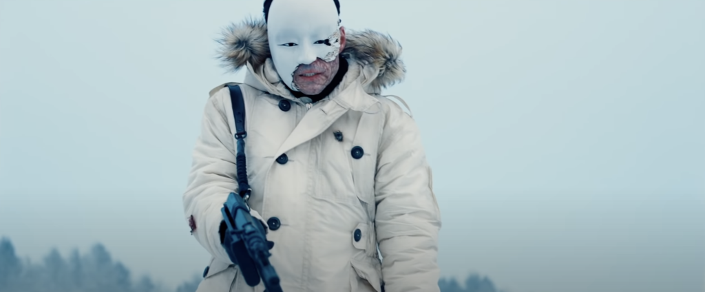 Shot of villain Safin, wearing a Japanese-style mask, holding a gun against a white backdrop.